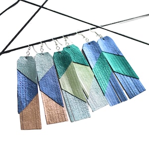 Pode incluir: Cinco pares de brincos de couro com formas geométricas e franjas. Os brincos são em tons de azul, verde e dourado.