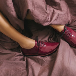 K&ouml;nnte beinhalten: Burgunderfarbene Lackschuhe mit schwarzen Schn&uuml;rsenkeln und spitzer Zehenpartie. Die Schuhe liegen auf einem Bett mit einer mauvefarbenen Bettdecke. Die Schuhe haben eine gen&auml;hte Zierleiste.