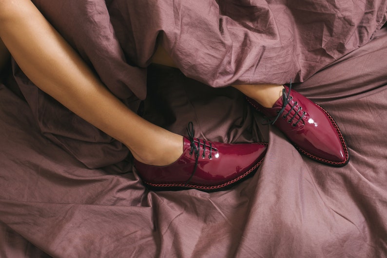 K&ouml;nnte beinhalten: Burgunderfarbene Lackschuhe mit schwarzen Schn&uuml;rsenkeln und spitzer Zehenpartie. Die Schuhe liegen auf einem Bett mit einer mauvefarbenen Bettdecke. Die Schuhe haben eine gen&auml;hte Zierleiste.