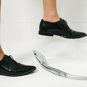 K&ouml;nnte beinhalten: Schwarze Lackschuhe mit spitzer Zehenpartie und dekorativer silberner Verzierung. Die Schuhe befinden sich in einem Badezimmer mit einem Duschkopf, der Wasser spr&uuml;ht. Die Schuhe stehen auf einer wei&szlig;en Oberfl&auml;che.
