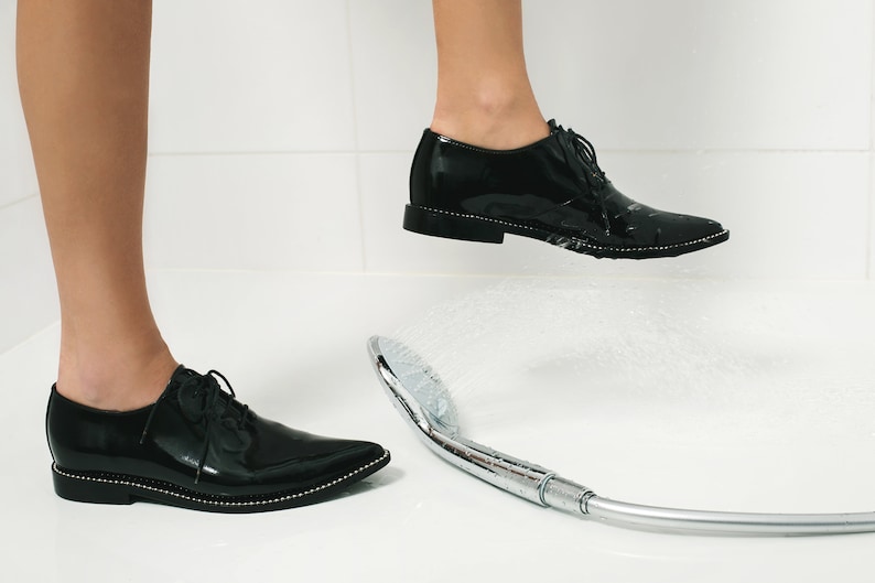 K&ouml;nnte beinhalten: Schwarze Lackschuhe mit spitzer Zehenpartie und dekorativer silberner Verzierung. Die Schuhe befinden sich in einem Badezimmer mit einem Duschkopf, der Wasser spr&uuml;ht. Die Schuhe stehen auf einer wei&szlig;en Oberfl&auml;che.