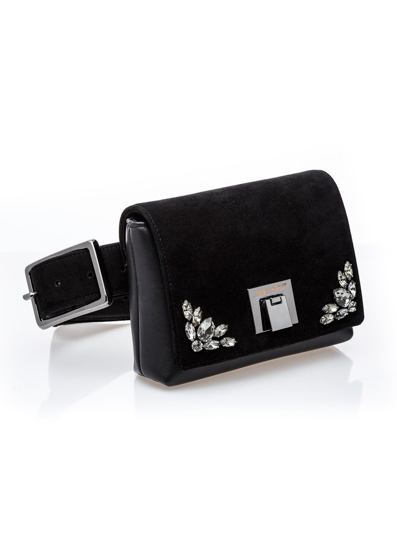 May include: Black suede belt bag with a silver buckle and crystal embellishments. The bag has a small flap with the brand name "Jimmy Choo" embossed on it.