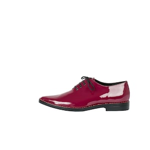 K&ouml;nnte beinhalten: Ein einzelner, gl&auml;nzender, weinroter Lederschuh mit spitzer Zehenpartie. Der Schuh hat schwarze Schn&uuml;rsenkel, eine schwarze Sohle und eine dekorative rote Naht am Rand der Sohle. Der Schuh ist vor wei&szlig;em Hintergrund platziert.