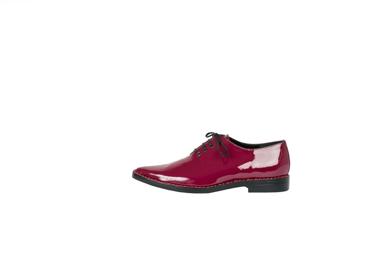 K&ouml;nnte beinhalten: Ein einzelner, gl&auml;nzender, weinroter Lederschuh mit spitzer Zehenpartie. Der Schuh hat schwarze Schn&uuml;rsenkel, eine schwarze Sohle und eine dekorative rote Naht am Rand der Sohle. Der Schuh ist vor wei&szlig;em Hintergrund platziert.