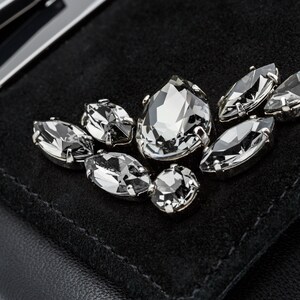 May include: A black suede clutch bag with a silver metal clasp and a decorative cluster of clear crystal rhinestones.