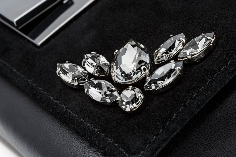 May include: A black suede clutch bag with a silver metal clasp and a decorative cluster of clear crystal rhinestones.