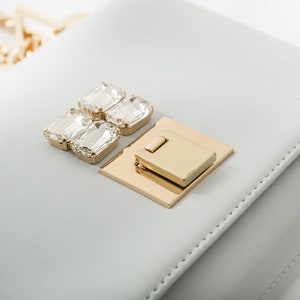 May include: A light gray leather handbag with a gold clasp and crystal embellishments.