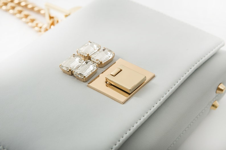 May include: A light gray leather handbag with a gold clasp and crystal embellishments.