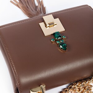 May include: A brown leather handbag with a gold chain strap and a gold clasp decorated with green gemstones. The bag has a brown tassel.