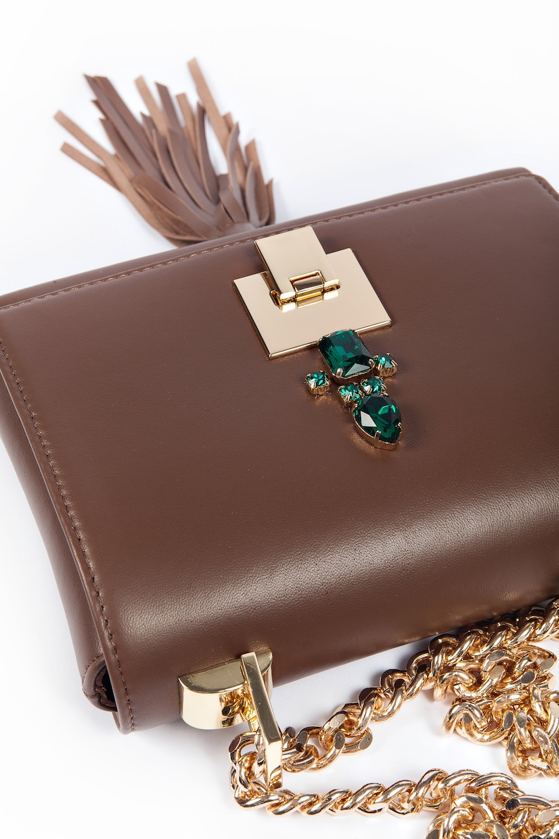 May include: A brown leather handbag with a gold chain strap and a gold clasp decorated with green gemstones. The bag has a brown tassel.