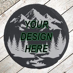 May include: Round black slate coaster with a mountain and forest design in white. The text "YOUR DESIGN HERE" is in green and black. The coaster is on a wooden surface.