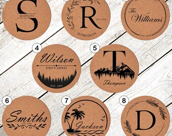 May include: A collection of eight round cork coasters with personalized designs. Each coaster features a different initial, name, or family name in black lettering, along with decorative elements like foliage, mountains, or a beach scene.