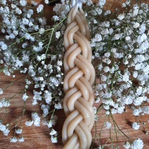 Op de afbeelding: Een gevlochten kaars van beige was met witte lonten. De kaars ligt op een houten oppervlak met witte bloemen.