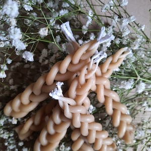 Op de afbeelding: Een set van vier gevlochten kaarsen met witte lonten. De kaarsen zijn lichtbruin van kleur en zijn in een kruispatroon gerangschikt. De kaarsen zijn omgeven door witte baby's breath bloemen.