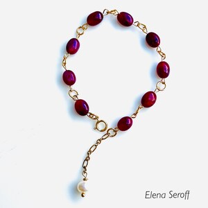 May include: A gold chain bracelet with a single white pearl charm and multiple dark red oval beads.