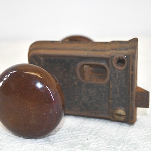 May include: An antique door knob and latch set. The knob is a glossy, oval shape in a rich brown color. The latch is rectangular, with a dark, aged finish, showing signs of rust.