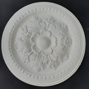 May include: A white, circular ceiling rose with an ornate floral and leaf pattern. The central design is a large flower, surrounded by smaller flowers and leaves. The outer edge has a beaded border.