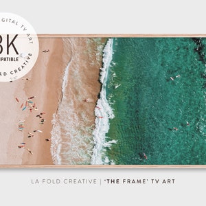 Könnte beinhalten: Luftaufnahme eines Strandes mit Surfern im türkisfarbenen Wasser und bunten Surfbrettern am Sand. Das Bild ist in einem hellen Holzrahmen gerahmt. Der Text auf dem Bild lautet "8K Compatible" und "LA FOLD CREATIVE | 'THE FRAME' TV ART."