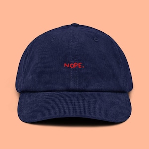 Può includere: Cappellino da baseball in velluto a coste blu navy con la scritta "NOPE" ricamata in rosso. Il cappellino ha una visiera curva e una corona strutturata. Design di Shive Design.