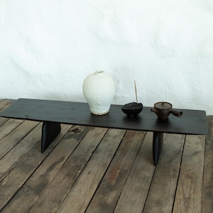 Puede incluir: Una mesa baja rectangular de madera negra con dos patas gruesas e inclinadas. Sobre la superficie hay un jarrón de cerámica de color blanco roto con textura, un cuenco pequeño oscuro con una varilla de incienso y una tetera de cerámica.