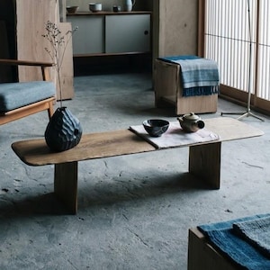 Puede incluir: Un sillón de madera con cojines de tela azul, una mesa de centro rectangular baja de madera, un jarrón negro texturizado, un pequeño cuenco de cerámica, una tetera, un taburete de madera con un paño azul y una lámpara de pie en una habitación con paneles de pared de madera.
