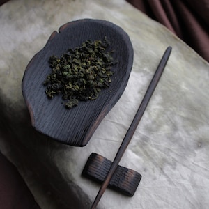 May include: A dark wooden tea scoop filled with green tea leaves, a long wooden tea spoon, and a small wooden spoon rest. The tea set is on a light-colored surface.