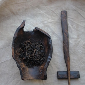 May include: A dark wooden tea bowl filled with loose tea leaves, next to a wooden tea pick and rest. The bowl has a unique, organic shape. The tea pick is long and slender, with a pointed end. The items are on a neutral-colored cloth.