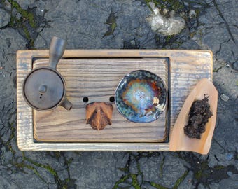 以下が含まれることがあります： 木製の長方形の茶盤の上に、ダークブラウンの陶器製横手急須、小さな茶こし、青とオレンジの色調の多色釉薬の茶碗、そして茶葉が入った薄茶色の茶さじが置かれています。