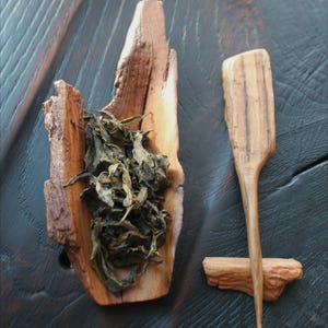 May include: A wooden tea scoop and a wooden tea bowl filled with dried tea leaves. The tea bowl is made from a piece of natural wood. The tea scoop has a long handle and a flat, paddle-like head. The items are on a dark wooden surface.