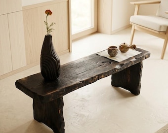 May include: A dark brown, charred wood bench with a live edge, featuring a tall, textured black vase holding a single orange flower, and a small ceramic cup and teapot resting on a white cloth square placed on the bench surface.