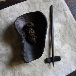 May include: A dark, matte black ceramic bowl with an irregular, organic shape containing small, dark, textured pieces. Beside the bowl lies a long, slender, pointed black wooden stick resting on a small, rectangular black ceramic rest.