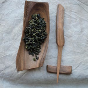 May include: A wooden tea scoop and tea tool set. The scoop is filled with loose leaf tea. The tea tool is a long, thin piece of wood with a handle and a stand. The set is made of natural wood.