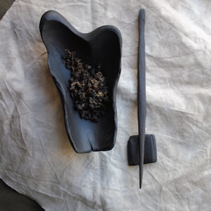 May include: A black wooden tea scoop and a tea tool rest on a white cloth. The scoop is filled with loose tea leaves. The tea tool is long and thin, with a small square base.