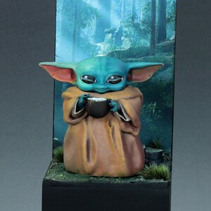 Op de afbeelding: Een klein, groen en blauw beeldje van een personage uit de Star Wars-franchise, dat een kopje vasthoudt, wordt getoond op een zwarte basis met een bosachtergrond.