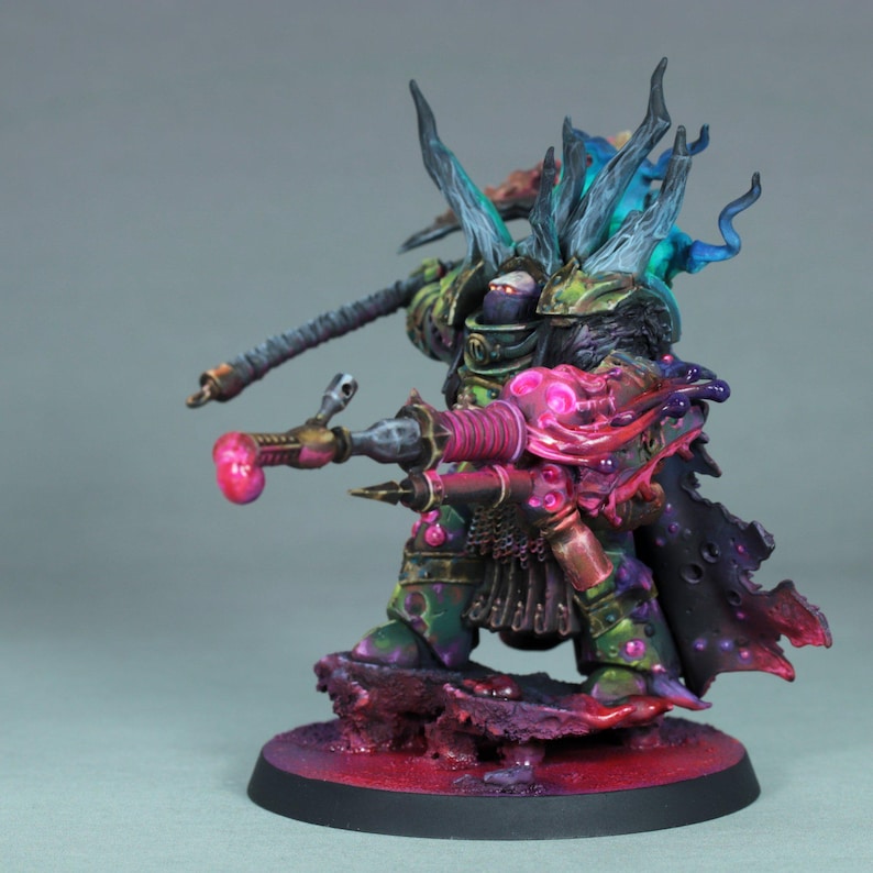May include: A detailed miniature figure of a warrior in green and bronze armor, holding a weapon. The figure has a helmet and is standing on a textured base with purple and red hues. The figure has a weapon with a pink tip.