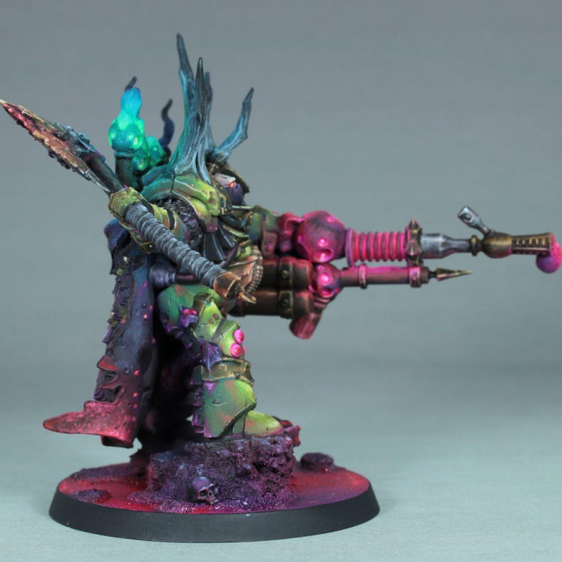 May include: A miniature figurine of a warrior in green, bronze, and purple armor, holding a weapon with a pink and bronze barrel. The figure has a horned helmet and a cape with red and purple accents. The base is dark with purple and red tones.