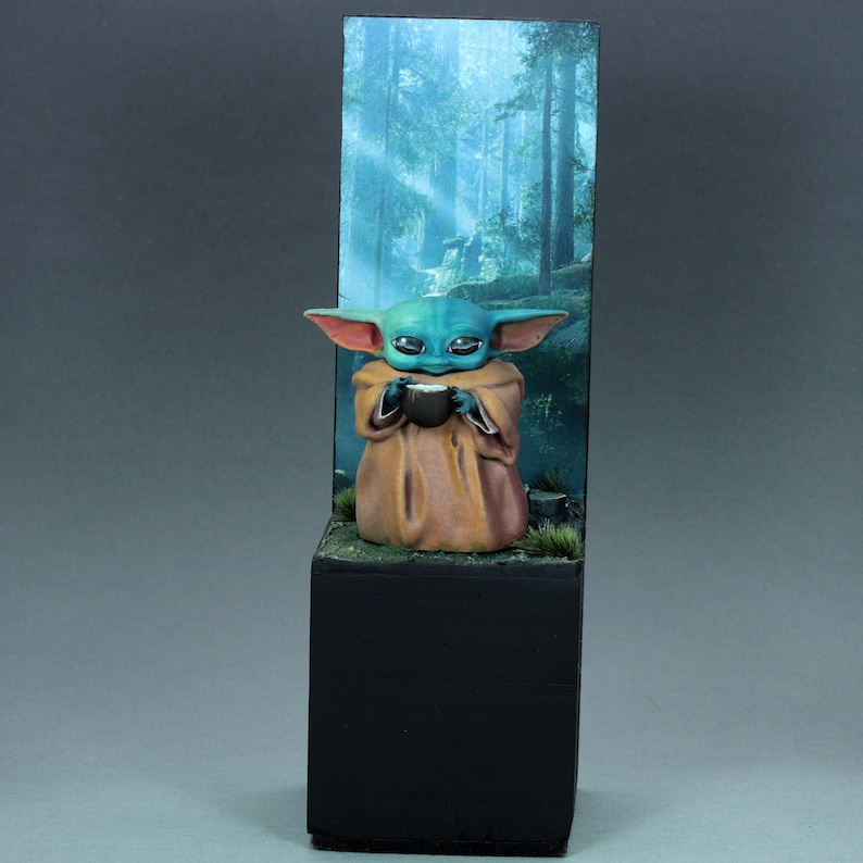 Op de afbeelding: Een klein, groen en blauw beeldje van een personage uit de Star Wars-franchise, dat een kopje vasthoudt, wordt getoond op een zwarte basis met een bosachtergrond.