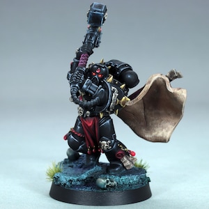 May include: A miniature figurine of a warrior in black armour, holding a large hammer. The figure has red accents, a red loincloth, and a tan cape. The base is dark blue with skulls and green grass.