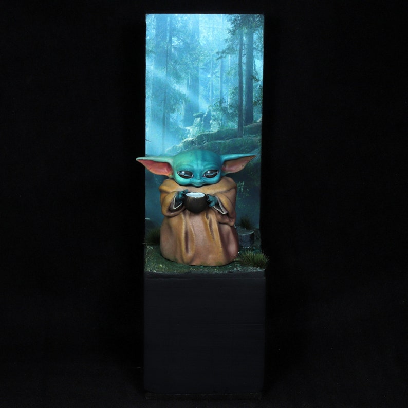 Op de afbeelding: Een klein groen en blauw beeldje van een personage uit de Star Wars-franchise, dat een klein kommetje vasthoudt. Het beeldje staat op een voetstuk met een groene en bruine grastextuur. De achtergrond is een wazig beeld van een bos.