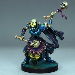 Puede incluir: Una miniatura pintada de una figura esquelética que lleva una armadura azul y morada, sosteniendo un bastón de hueso y un tambor con forma de cráneo. La figura tiene ojos amarillos brillantes y una expresión amenazante.