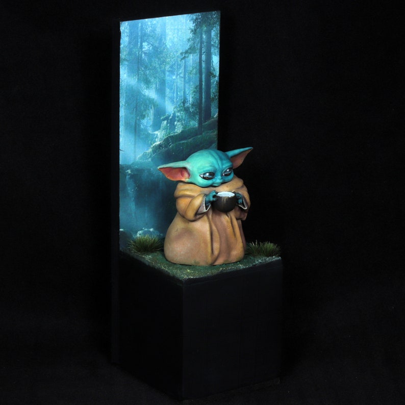 Op de afbeelding: Een klein groen en blauw beeldje van een personage uit de Star Wars-franchise, zittend op een zwarte basis met een bosachtergrond. Het personage houdt een klein kopje in zijn handen.