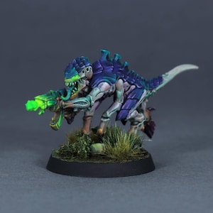 Könnte beinhalten: Eine Miniatur-Alien-Kreatur-Figur mit einem violett-blauen gepanzerten Rücken, blasser Haut und einem langen Schwanz. Die Kreatur hat ein breites, zahniges Maul und hält eine leuchtend neongrüne Waffe. Sie steht auf einem runden schwarzen Sockel.