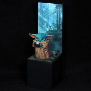 Op de afbeelding: Een klein, groen en blauw beeldje van een personage uit de Star Wars-franchise, dat een kopje vasthoudt, staand op een basis met een bosachtergrond.