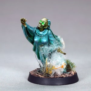 Puede incluir: Una figura miniatura pintada de un personaje fantasmal con ojos verdes brillantes, que lleva una túnica azul fluida y está rodeado de telarañas blancas. La figura está representada con un brazo levantado y el otro extendido, como si estuviera alcanzando algo. La figura está colocada sobre una pequeña base redonda con tierra marrón y hierba verde.