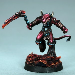 Puede incluir: Una figurita miniatura pintada de rojo y azul de un demonio con cuernos y cola, sosteniendo un arma roja y azul. La figurita está de pie sobre una base de rocas pintadas de rojo y negro.