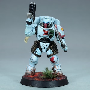 May include: A miniature figurine of a space marine in light blue armor, holding a black pistol and a silver blade. The figurine has a red skull emblem on its chest and a red face mask. It stands on a base with a red and brown surface.
