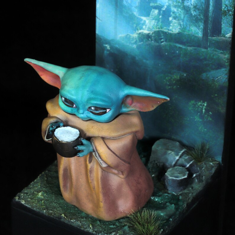 Op de afbeelding: Een klein, groen en blauw beeldje van een personage uit de Star Wars-franchise, dat een klein kopje vasthoudt. Het beeldje draagt een bruine robe en staat op een donkerbruine ondergrond met groen gebladerte.