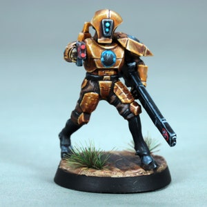 Puede incluir: Una figura en miniatura pintada de oro de un soldado que lleva un casco con una visera azul y sostiene un rifle negro. La figura está de pie sobre una base redonda con pintura texturizada marrón y verde.