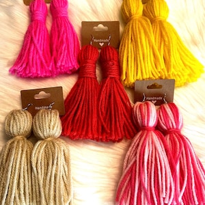 May include: A collection of handmade tassel earrings in various colors, including hot pink, yellow, red, beige, and pink and white. Each pair is attached to a brown card with the word "Handmade".