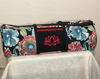 May include: A cylindrical duffel bag featuring a black background with a vibrant floral pattern in shades of red, teal, and white. The bag includes a solid black center panel with a red lotus flower graphic and a red zippered pocket.